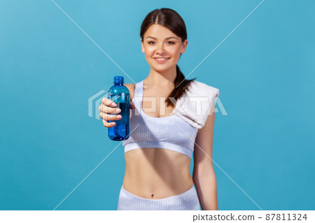 Healthy smiling woman athlete in white sportswear with towel on shoulder holding and showing blue bottle with mineral water, restoring electrolytes. Indoor studio shot isolated on blue background 87811324
