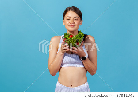 Athletic slim woman in white sports top closing eyes enjoying aroma of fresh vegetable salad in big bowl, healthy nutrition, detox diet. Indoor studio shot isolated on blue background 87811343