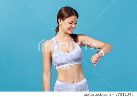 Positive sports woman in white tights and top looking at smartwatch on her wrist, monitoring her indicators after workout, satisfied with results. Indoor studio shot isolated on blue background 87811345