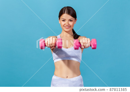 Smiling healthy athletic woman in sportswear holding dumbbells in hands, showing her abs, doing exercises to keep good body shape. Indoor studio shot isolated on blue background Smiling healthy athletic woman in sportswear holding dumbbells in hands, showing her abs, doing exercises to keep good body shape. Indoor studio shot isolated on blue background 87811385