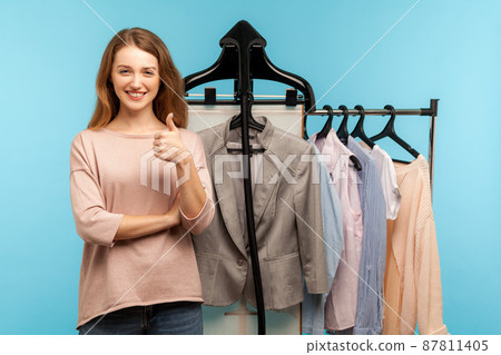 Optimistic woman, fashion stylist standing near business clothes hang on shelf in designer store and showing thumbs up, selling trendy outfit collection in shop boutique. indoor studio shot isolated Optimistic woman, fashion stylist standing near business clothes hang on shelf in designer store and showing thumbs up, selling trendy outfit collection in shop boutique. indoor studio shot isolated 87811405