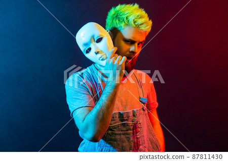 Portrait of handsome hipster man with stylish haircut holding white mask, standing with raised eyebrow and amazed face, multiple personality disorder. Colorful neon light, indoor studio shot. 87811430