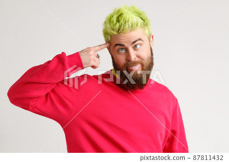Handsome bearded funny guy holding pistol figure with fingers to temple, crossing eyes like squint, tired from routine, wearing red sweater. Indoor studio shot isolated on gray background. 87811432