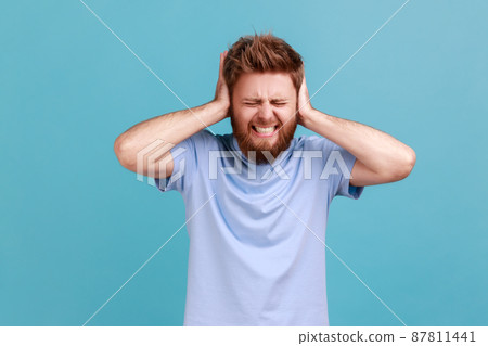 Don't want to listen. Portrait of despaired man irritated by loud noise covering ears and grimacing in pain, suffering annoying high-decibel sound. Indoor studio shot isolated on blue background. 87811441