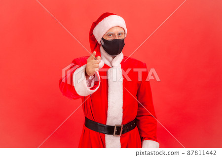 Serious santa claus in medical protective mask pointing finger at camera, warning about epidemic situation, covid-19 prevention. Indoor studio shot isolated on red background 87811442