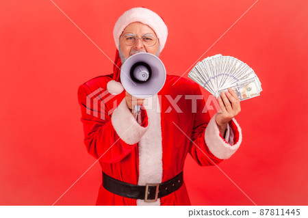 Elderly gray bearded santa claus screaming in megaphone holding fan of dollars, holidays bonuses and promotions. Indoor studio shot isolated on red background 87811445