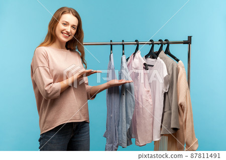 Lovely woman, fashion stylist pointing clothes hang on shelf in designer store, showing wardrobe with trendy outfit on rack in modern shop boutique. indoor studio shot isolated on blue background Lovely woman, fashion stylist pointing clothes hang on shelf in designer store, showing wardrobe with trendy outfit on rack in modern shop boutique. indoor studio shot isolated on blue background 87811491