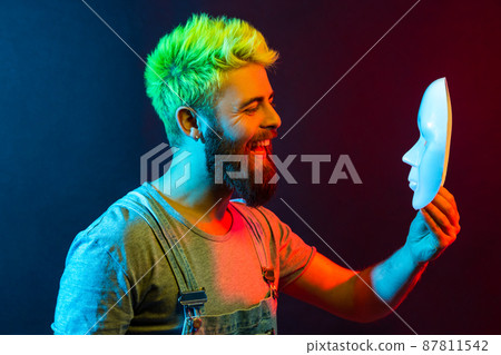 Happy bearded hipster man holding and looking at white mask with toothy smile, being satisfied of his hidden personality, expressing joy and happiness. Colorful neon light, indoor studio shot. Happy bearded hipster man holding and looking at white mask with toothy smile, being satisfied of his hidden personality, expressing joy and happiness. Colorful neon light, indoor studio shot. 87811542