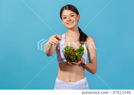 Smiling woman is white sportswear with slim body pointing finger on you holding bowl with vegetable salad, suggesting nutritional diet. Indoor studio shot isolated on blue background 87811546