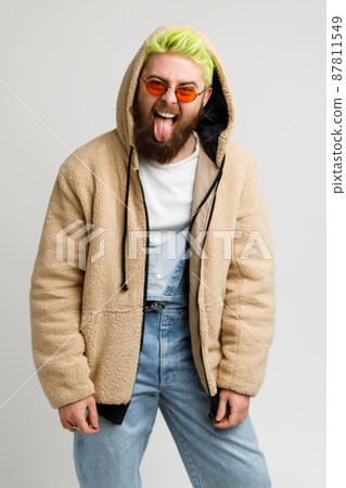 Handsome bearded man wears in stylish jacket and denim overalls fashion hair style looking at camera with crazy expression, showing tongue. Indoor studio shot isolated over gray background. Handsome bearded man wears in stylish jacket and denim overalls fashion hair style looking at camera with crazy expression, showing tongue. Indoor studio shot isolated over gray background. 87811549