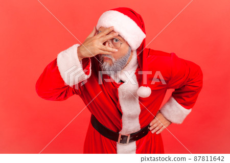 Portrait of curious nosy man in santa claus costume pretending to close eyes with hand attentively peeping through fingers, spying. Indoor studio shot isolated on red background 87811642