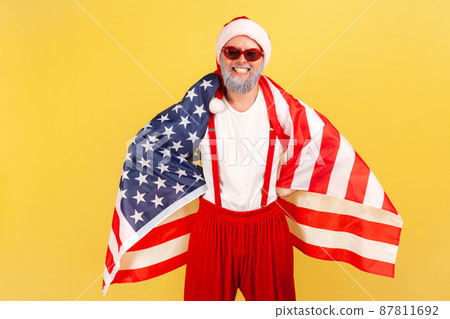Happy patriotic grey bearded man in santa claus hat and sunglasses standing wrapped in usa flag and looking at camera with toothy smile. Indoor studio shot isolated on yellow background 87811692