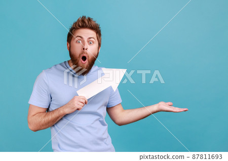 Portrait of bearded man holding arrow pointing up looking at camera with open mouth, growth and increase concept, presenting copy space. Indoor studio shot isolated on blue background. 87811693