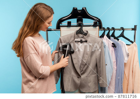 Cheerful housewife steaming business jacket at dry-cleaner and looking at fabric, advertisement of professional services washing, cleaning and ironing garment. indoor studio shot, blue background Cheerful housewife steaming business jacket at dry-cleaner and looking at fabric, advertisement of professional services washing, cleaning and ironing garment. indoor studio shot, blue background 87811716