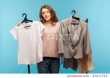 Uncertain confused woman shopper holding clothes on racks and looking with doubts, choosing between casual shirt and classic business jacket, shopping in modern boutique. indoor studio shot isolated 87811755