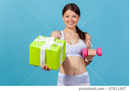 Happy athletic woman in white sportswear holding green gift box and pink dumbbell in hand, preparing sports equipment as presents on holidays. Indoor studio shot isolated on blue background Happy athletic woman in white sportswear holding green gift box and pink dumbbell in hand, preparing sports equipment as presents on holidays. Indoor studio shot isolated on blue background 87811807
