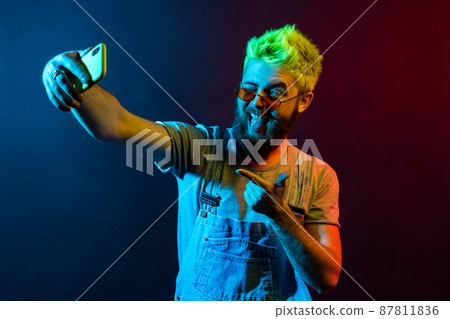 Handsome hipster bearded man wearing t shirt and denim overalls, broadcasting live stream, points to camera with finger, grimacing, winking and showing tongue. Colorful neon light, indoor studio shot 87811836