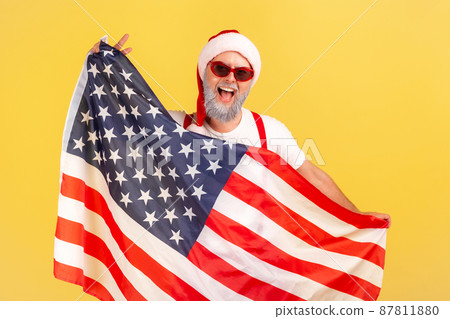 Patriotic happy gray bearded man in stylish sunglasses and santa claus hat holding flag of united states of America, voting on winter election. Indoor studio shot isolated on yellow background 87811880