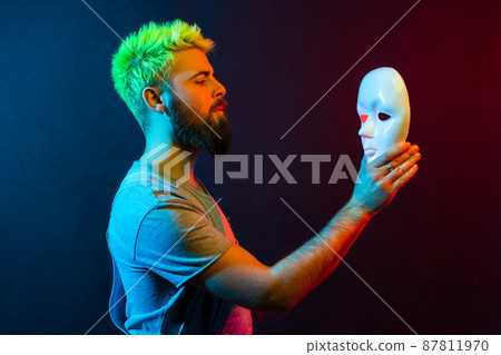 Side view of confident serious man in denim overalls, holding and looking at white mask in hands with attentive look, trying to understand hiding personality. Colorful neon light, indoor studio shot. Side view of confident serious man in denim overalls, holding and looking at white mask in hands with attentive look, trying to understand hiding personality. Colorful neon light, indoor studio shot. 87811970