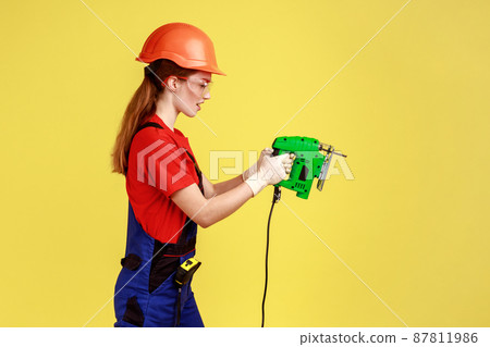 Side view portrait of builder woman working with fretsaw, expressing serious emotions and confidence, wearing overalls and protective helmet. Indoor studio shot isolated on yellow background. Side view portrait of builder woman working with fretsaw, expressing serious emotions and confidence, wearing overalls and protective helmet. Indoor studio shot isolated on yellow background. 87811986