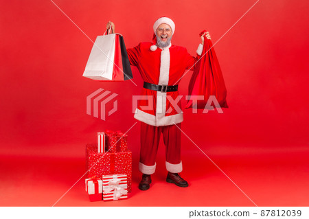 Full length happy satisfied santa claus holding and showing paper bags and big red bag full of presents, gift boxes standing on floor, winter holidays. Indoor studio shot isolated on red background Full length happy satisfied santa claus holding and showing paper bags and big red bag full of presents, gift boxes standing on floor, winter holidays. Indoor studio shot isolated on red background 87812039
