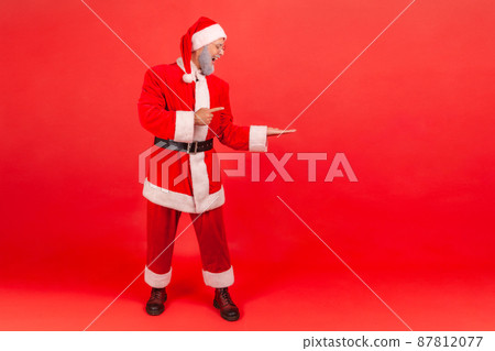 Full length portrait of positive happy man in santa claus clothes pointing finger away showing empty space for advertisement, freespace. Indoor studio shot isolated on red background 87812077