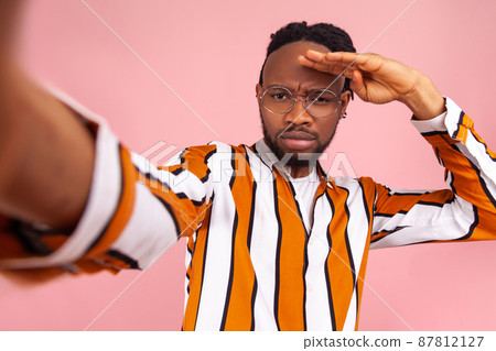 Serious assertive african man blogger with dreadlocks, in striped shirt looking far away with hand over head, looking for new subscribers, posing selfie. Indoor studio shot isolated on pink background 87812127