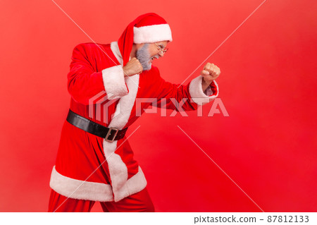 Side view portrait of aggressive purposeful gray bearded man holding fists up, fighting with competitors, pretending to be santa claus, fight spirit on holidays. Indoor isolated on red background Side view portrait of aggressive purposeful gray bearded man holding fists up, fighting with competitors, pretending to be santa claus, fight spirit on holidays. Indoor isolated on red background 87812133