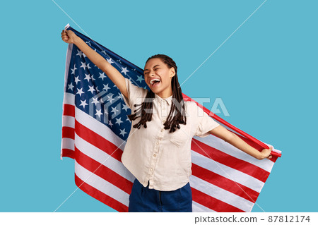 Cheerful attractive woman with black dreadlocks holding USA flag over shoulders and keeps eyes closed and smiling happily, wearing white shirt. Indoor studio shot isolated on blue background. Cheerful attractive woman with black dreadlocks holding USA flag over shoulders and keeps eyes closed and smiling happily, wearing white shirt. Indoor studio shot isolated on blue background. 87812174