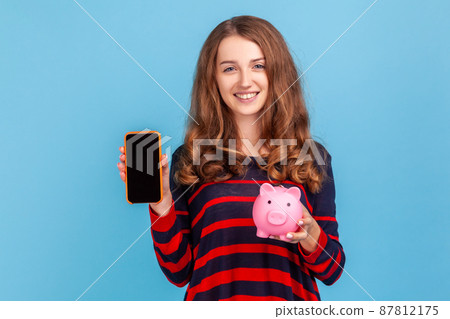 Positive woman wearing striped casual style sweater, holding piggy bank for saving money and smartphone with blank screen for advertisement. Indoor studio shot isolated on blue background. Positive woman wearing striped casual style sweater, holding piggy bank for saving money and smartphone with blank screen for advertisement. Indoor studio shot isolated on blue background. 87812175