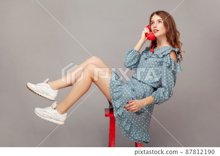 Charming girl in ruffle dress sitting delicate graceful airy on chair, holding phone handset enjoying pleasant conversation on telephone, looking up dreamy relaxed. indoor studio shot isolated on gray Charming girl in ruffle dress sitting delicate graceful airy on chair, holding phone handset enjoying pleasant conversation on telephone, looking up dreamy relaxed. indoor studio shot isolated on gray 87812190