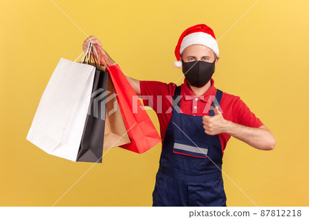 Post man in santa claus hat and protective medical mask on face showing thumbs up holding shopping bags, goods delivery during quarantine. Indoor studio shot isolated on yellow background Post man in santa claus hat and protective medical mask on face showing thumbs up holding shopping bags, goods delivery during quarantine. Indoor studio shot isolated on yellow background 87812218