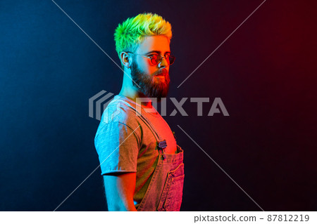 Side view portrait of guy with beard and green hair, looking at camera with serious and confident expression, wearing denim overalls, copy space. Colorful neon light, indoor studio shot. Side view portrait of guy with beard and green hair, looking at camera with serious and confident expression, wearing denim overalls, copy space. Colorful neon light, indoor studio shot. 87812219