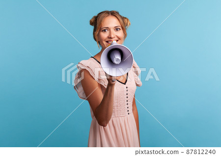 Young adult blonde woman in summer dress talking with megaphone, proclaiming news, loudly announcing advertisement, warning using loudspeaker. Indoor studio shot isolated on blue background. 87812240
