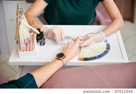 Crop view of manicure specialist filing woman nails with nail file. Female manicurist in sterile gloves doing manicure for client in beauty salon. 87812848