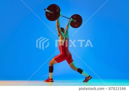 Studio shot of young man in red sportswear exercising with barbell isolated blue background in neon. Sport, weightlifting, power, achievements concept 87813166