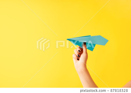 A woman's hand holds blue paper airplane on yellow background A woman's hand holds blue paper airplane on yellow background 87818328