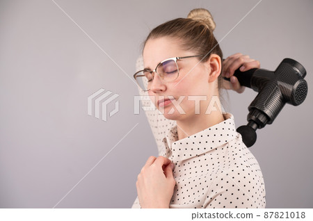 Caucasian business lady makes herself a back massage with a massager gun on a white background. Caucasian business lady makes herself a back massage with a massager gun on a white background. 87821018
