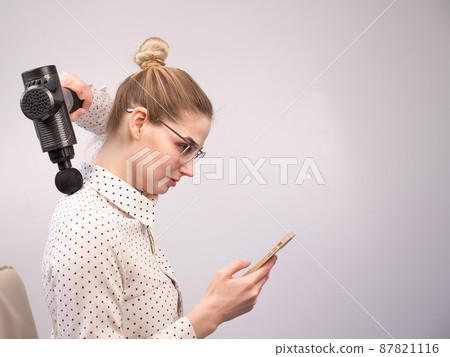 A young woman sits on a chair, uses a smartphone and makes herself massage with an electro massage gun. A young woman sits on a chair, uses a smartphone and makes herself massage with an electro massage gun. 87821116
