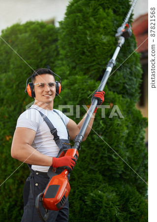 Positive caucasian man with petrol hedge trimmer in hands standing on back yard and looking at camera. Competent gardener trimming plants on fresh air. 87821568