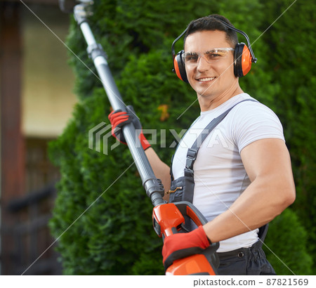 Portrait of strong caucasian man in overalls, safety glasses, headphones and gloves posing at garden with electric hedge trimmer in hands. Pruning process during summer time. 87821569