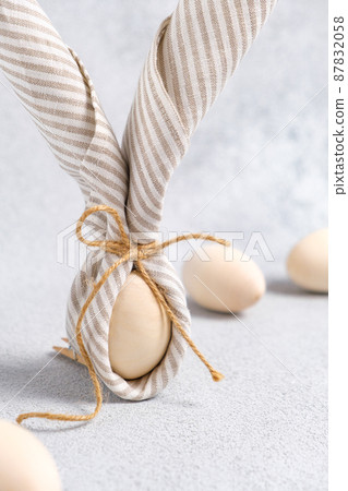 Close-up of Easter bunny made from egg and linen striped napkin. Easter eggs are light beige on light gray background. Trend of decorating traditional spring holiday Easter. Vertical, pastel colors. 87832058