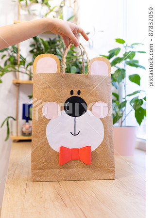 Paper bag in form of teddy bear head with black nose and bow tie in child's hand against background of houseplants. Shopping, birthday present or gift. Eco-friendly packing. DIY. 87832059