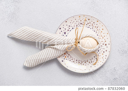 Egg wrapped in linen striped napkin tied with jute in form Easter bunny with ears form on plate, light gray background. Idea of Easter table setting in pastel colors. 87832060