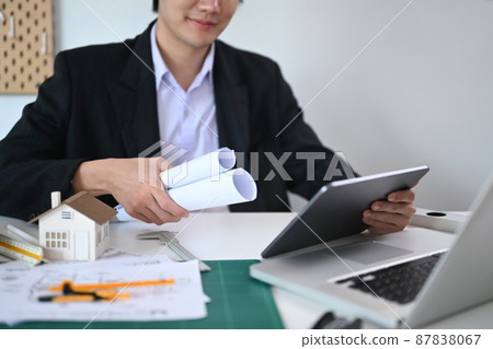 Architect male holding blueprint roll and working on architectural projects in office. Architect male holding blueprint roll and working on architectural projects in office. 87838067