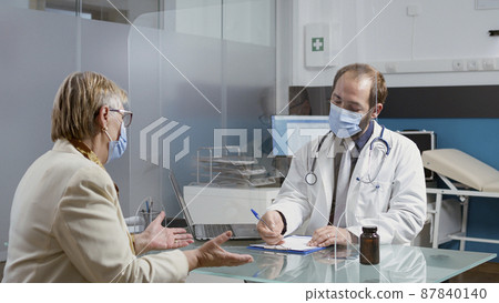 Specialist taking notes at consultation with senior patient, writing prescription treatment papers during pandemic. Medic and elder woman talking about healthcare service in cabinet. 87840140