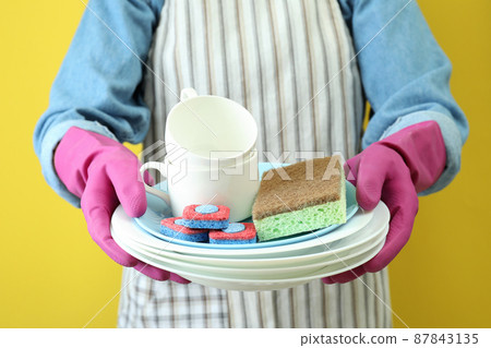 Woman in apron holds clean dishware on yellow background 87843135
