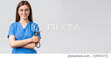 Healthcare workers, medicine, insurance and covid-19 pandemic concept. Confident and enthusiastic young female nurse, doctor in blue scrubs and glasses, hold stethoscope, smiling camera pleased 87845731