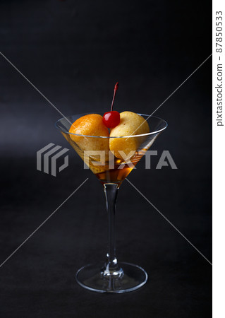 Neapolitan Rum baba (or baba au rum) in a martini glass with a cocktail cherry on a black background. Small yeast cakes soaked in rum syrup. Traditional Italian pastry. Close-up, selective focus. Neapolitan Rum baba (or baba au rum) in a martini glass with a cocktail cherry on a black background. Small yeast cakes soaked in rum syrup. Traditional Italian pastry. Close-up, selective focus. 87850533