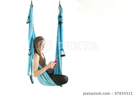 Yoga instructor has break and holding phone. Young athletic woman hang in hammock during an aerial gymnastics or fly yoga workout. Yoga instructor has break and holding phone. Young athletic woman hang in hammock during an aerial gymnastics or fly yoga workout. 87854331